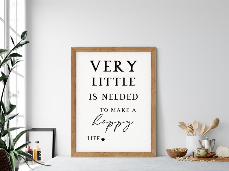 May include: A framed print with the text "VERY LITTLE IS NEEDED TO MAKE A happy LIFE" in black on a white background. The frame is made of light brown wood. The print is displayed on a table with kitchen utensils and decor.