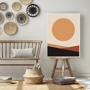 May include: A framed abstract art print featuring a large orange sun over a landscape of brown and black geometric shapes. The artwork is displayed in a room with a bench, woven baskets, and decorative wall hangings, creating a warm, inviting atmosphere.