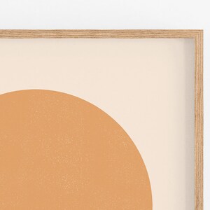 May include: A framed abstract art print featuring a large, textured, orange-brown semi-circle against a cream-colored background. The artwork is encased in a light-colored wooden frame, creating a minimalist aesthetic.