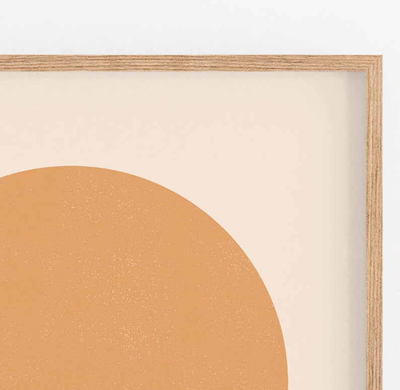 May include: A framed abstract art print featuring a large, textured, orange-brown semi-circle against a cream-colored background. The artwork is encased in a light-colored wooden frame, creating a minimalist aesthetic.
