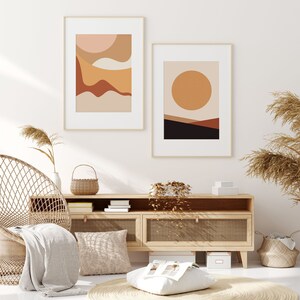 May include: Two framed abstract art prints with gold frames. One print features layered shapes in peach, tan, and brown. The other print has a large orange circle over a landscape of brown and black. The prints hang above a wooden cabinet.