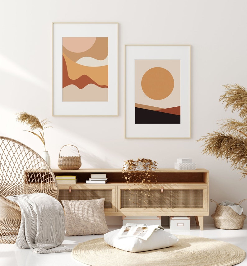 May include: Two framed abstract art prints with gold frames. One print features layered shapes in peach, tan, and brown. The other print has a large orange circle over a landscape of brown and black. The prints hang above a wooden cabinet.