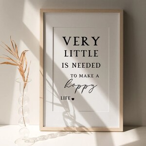 May include: Framed print with the text "VERY LITTLE IS NEEDED TO MAKE A happy LIFE" in black font. The print is in a light wood frame with a white mat. A clear glass vase with a dried plant is in the foreground.