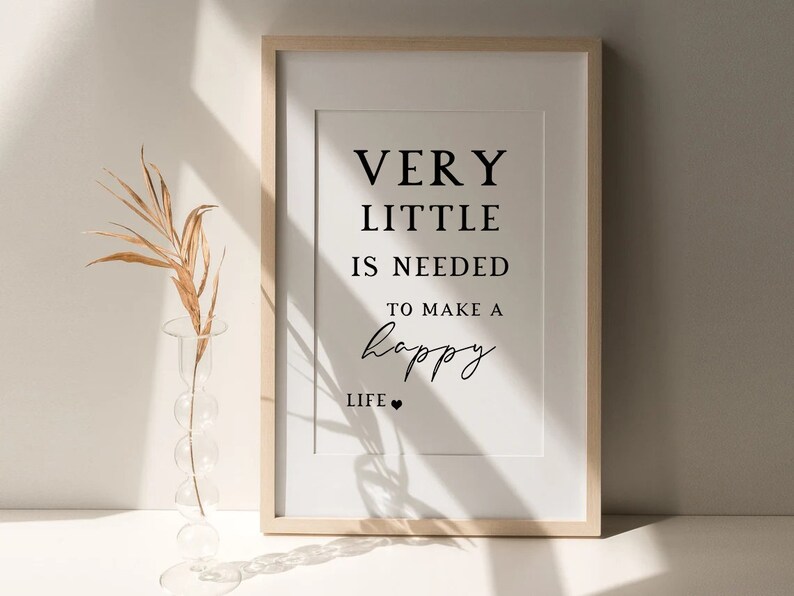 May include: Framed print with the text "VERY LITTLE IS NEEDED TO MAKE A happy LIFE" in black font. The print is in a light wood frame with a white mat. A clear glass vase with a dried plant is in the foreground.