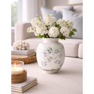 May include: A white ceramic vase with a rounded body and a narrow neck, featuring hand-painted blue floral motifs. The vase is filled with a large, dense bouquet of white lilac flowers with green leaves, creating a bright and floral arrangement.