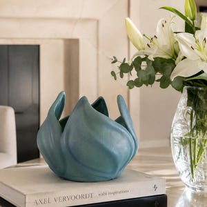 May include: A matte teal ceramic bowl with a sculptural, organic shape resembling an opening flower bud. The bowl is positioned on top of two stacked hardcover books, one titled Axel Vervoordt wabi inspirations and the other Joseph Dirand Interior.