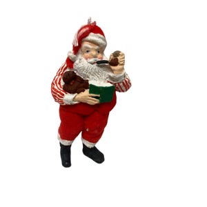 May include: A vintage Santa Claus figurine, dressed in a red suit with white trim and a matching hat. The figurine is holding a green book and a brown teddy bear, and smoking a pipe. A classic Christmas decoration.