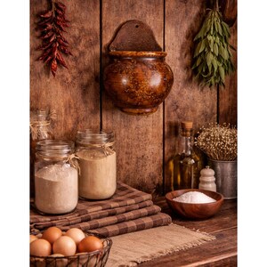 May include: A wall-mounted brown ceramic salt pig with a speckled finish, positioned above a wooden surface containing two glass jars filled with flour, a wooden bowl of salt, a wire basket of brown eggs, and a bottle of oil.