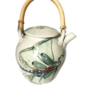 May include: A ceramic teapot with a bamboo handle and lid. The teapot is white with a hand-painted dragonfly design in shades of blue, green, and red. The handle is curved and attached to the teapot with small woven pieces.