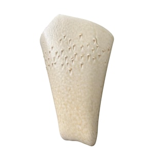 May include: A cream-colored, textured ceramic vase with a unique, asymmetrical shape. The vase features a series of small, triangular indentations near the top, adding a decorative element. The surface has a speckled appearance.