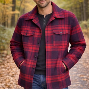 May include: A long-sleeve button-up shirt jacket featuring a red and dark purple plaid pattern, a pointed collar, two chest pockets with buttoned flaps, and two side pockets. The garment is worn over a black henley shirt and blue denim jeans.