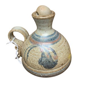 May include: A handmade ceramic pitcher with a handle and a lid. The pitcher is beige with blue and orange stripes around the neck and a dark blue and black design on the body. The pitcher has a rustic, speckled appearance.