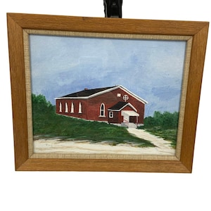 May include: A framed painting of a red brick building with a white entrance and a cross symbol. The building is set against a blue sky and green foliage, with a dirt path leading to it.
