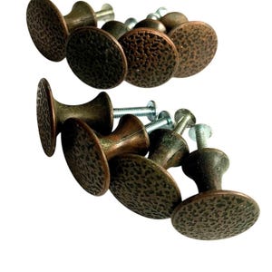 May include: A collection of bronze-colored cabinet knobs with a hammered texture. Each knob has a rounded face and a stem with a screw for installation. The knobs are arranged against a white background.