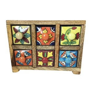 May include: A small wooden chest of drawers with six colorful ceramic tile fronts. Each drawer front features a unique floral or geometric design in shades of yellow, orange, blue, and green. The knobs are round and painted in complementary colors.