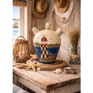May include: A decorative ceramic jug with a lighthouse design. The jug features a red and black lighthouse on a blue and white background. A woven lantern, starfish, and seashells are on a wooden table.