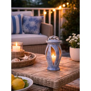 May include: A blue ceramic lantern with a rope handle and cut-out designs, illuminated by a lit candle. The lantern sits on a brown tile table, with a wicker tray, seashells, and a bowl of lemons nearby. A lit candle in a glass holder is also on the table.