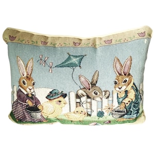 May include: Decorative Easter-themed pillow featuring a light blue background with a pastoral scene. The image depicts two rabbits, two chicks, and a kite near a white picket fence. The pillow has a beige border with floral accents.