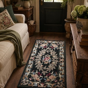 May include: A rectangular area rug featuring a black background with a central oval motif of light gray leaves and pink, blue, and cream floral patterns. The border displays a repeating intricate design of colorful flowers and foliage.