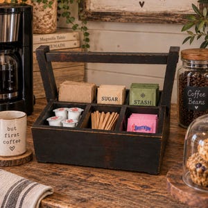 May include: A dark brown wooden caddy with a handle and six compartments, organized to hold various coffee and tea accessories. The compartments contain small white creamer cups, wooden stirrers, and packets labeled Sugar, Stash, and Sweet'N Low.