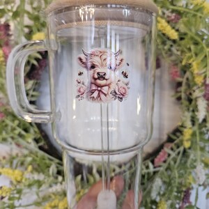 May include: A clear glass mug with a handle, a bamboo lid, and a glass straw. The mug features a watercolour illustration of a Highland cow with pink flowers and bees. The mug is designed for cold drinks.