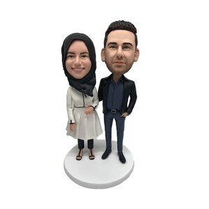 May include: A custom bobblehead figurine set featuring a couple. The woman is wearing a white coat and a black headscarf. The man is wearing a black jacket and blue jeans. The figurines are standing on a white base.