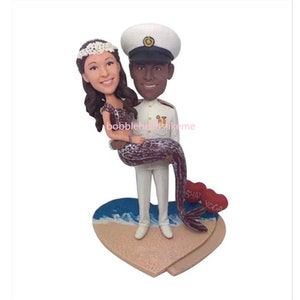 May include: A custom bobblehead figurine of a couple. The male figure is wearing a white naval uniform with a navy blue hat. The female figure is wearing a white dress with a purple and white mermaid tail. The couple is standing on a heart-shaped base with a beach scene and the words "Say" and "Yoga" on the hearts.