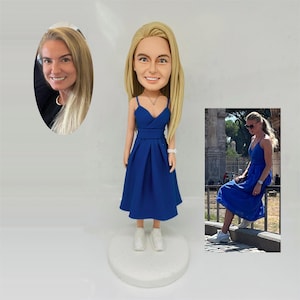 Op de afbeelding: Een op maat gemaakte bobblehead-figuur van een vrouw in een blauwe jurk, met een bijpassende foto. Het beeldje heeft blond haar en draagt een blauwe jurk met witte sneakers. De foto toont de vrouw in een vergelijkbare jurk.
