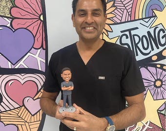 May include: A custom bobblehead figurine depicting a person with short dark hair, wearing a grey V-neck scrub top and matching trousers, standing with arms crossed on a circular grey base, held in front of a person wearing a black scrub top.