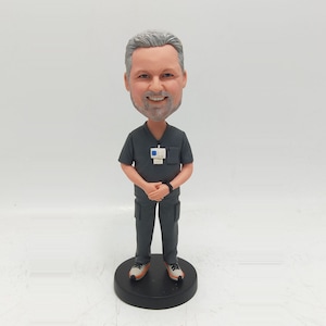 May include: A custom bobblehead figure of a person with grey hair and a beard, wearing a dark grey scrub top and matching trousers. The figure has a white identification badge clipped to the chest and is standing on a round black base.