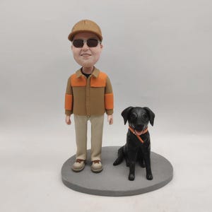 May include: A figurine of a man wearing sunglasses, a brown jacket with orange accents, and khaki pants, standing next to a black dog with an orange collar. The man is wearing a brown cap. Both figures are on a gray circular base.