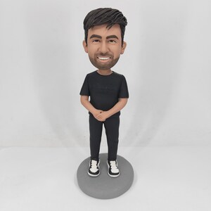 Op de afbeelding: Een op maat gemaakte bobblehead-figuur van een man in een zwart t-shirt, zwarte broek en sneakers. De figuur heeft een lachend gezicht en staat op een grijze, cirkelvormige basis. De bobblehead is een gepersonaliseerd cadeau.