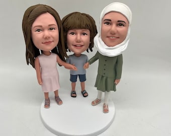 May include: Three custom-made figurines of a family. The figures are made of clay and painted in realistic detail. The woman is wearing a green dress and a white headscarf. The girl is wearing a pink dress and the boy is wearing a blue shirt and blue shorts.
