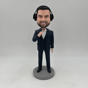 Puede incluir: Una figura de cabezón personalizada de una persona que lleva un traje azul oscuro, una camisa blanca y auriculares negros. La persona sostiene un micrófono en su mano derecha.