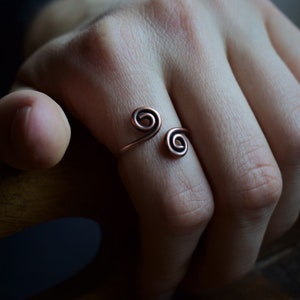 Könnte beinhalten: Ein kupferfarbener Ring mit einem doppelten Spiraldesign. Der Ring wird an einem Finger getragen und unterstreicht seinen minimalistischen und künstlerischen Stil. Das offene Design ermöglicht eine verstellbare Passform. Der warme Farbton des Rings ergänzt den Hautton.