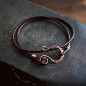 May include: A brown leather bracelet with a copper hook clasp. The hook is a simple, elegant design with a swirl at the top.