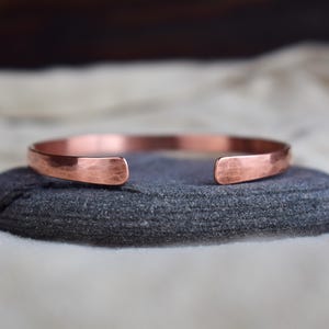 May include: A hammered copper bracelet with a simple, minimalist design. The bracelet is made from a single piece of copper that has been hammered to create a textured surface.