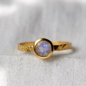 Puede incluir: Un anillo de oro con una piedra de luna redonda e iridiscente engastada en un engaste de bisel. La banda tiene un acabado texturizado martillado.