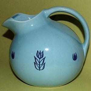 May include: A light blue ceramic pitcher with a rounded body and a curved handle. The pitcher features a spout for pouring and is decorated with three dark blue tulip designs. The pitcher is likely used for serving beverages.
