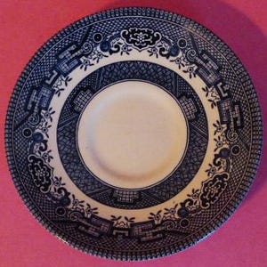 May include: A round, white and blue ceramic plate with a detailed, ornate pattern. The plate has a central white circle surrounded by a blue design. The outer rim of the plate is also blue, with a repeating pattern. The plate is set against a pink background.