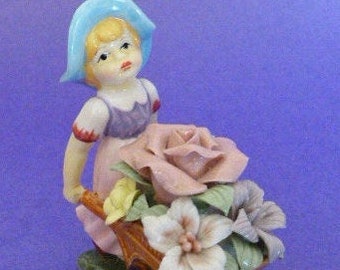 May include: A porcelain figurine of a girl wearing a blue hat and a purple dress, pushing a wheelbarrow filled with pink, white, and yellow flowers.