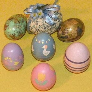 May include: Six decorated Easter eggs, including a blue egg with a white chick design, a purple egg with a floral design, a pink egg with a yellow flower design, a green egg with a brown speckled pattern, a gold egg with a brown pattern, and a white egg with a blue ribbon and floral design.