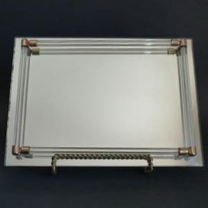 May include: Rectangular mirrored tray with clear acrylic and copper-toned accents. The tray has a decorative metal handle and is designed to be displayed on a stand. The frame is made of clear acrylic rods.