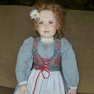 May include: A porcelain doll wearing a blue and white dress with a red and floral patterned vest. The doll has red shoes and a white flower in her hair.