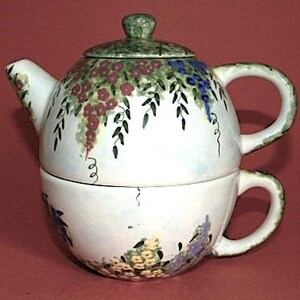 May include: A white ceramic teapot and teacup set with a floral design. The teapot has a green lid and handle, and the teacup has a matching handle. The set is decorated with purple, blue, and yellow flowers.