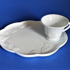 May include: A white ceramic teacup and saucer set. The saucer is oval-shaped with embossed grape and leaf designs. The teacup has a handle and is placed on the saucer. The set is on a blue background.