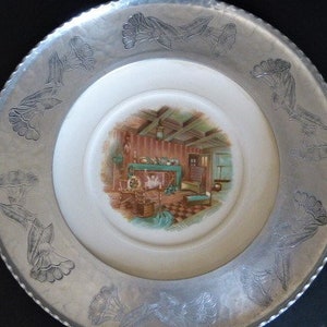 May include: A silver aluminum serving tray with a raised rim and a floral design. The center of the tray features a white porcelain insert with a painted scene of a room with a fireplace.