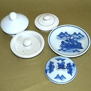 May include: A collection of small, white ceramic dishes and lids. One dish features a blue and white landscape design. The lids have a knob on top. The dishes are arranged on a light brown surface.