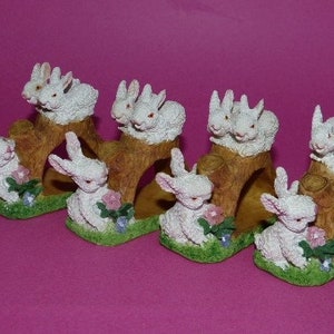 May include: Set of six white bunny napkin rings, each bunny is sitting on a brown tree stump with green grass and pink flowers.