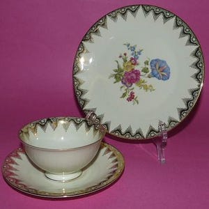 May include: A vintage tea set with a floral design. The set includes a teacup, saucer, and a plate. The teacup and saucer have gold trim and a black and gold patterned border. The plate features a floral bouquet with pink, blue, and yellow flowers.